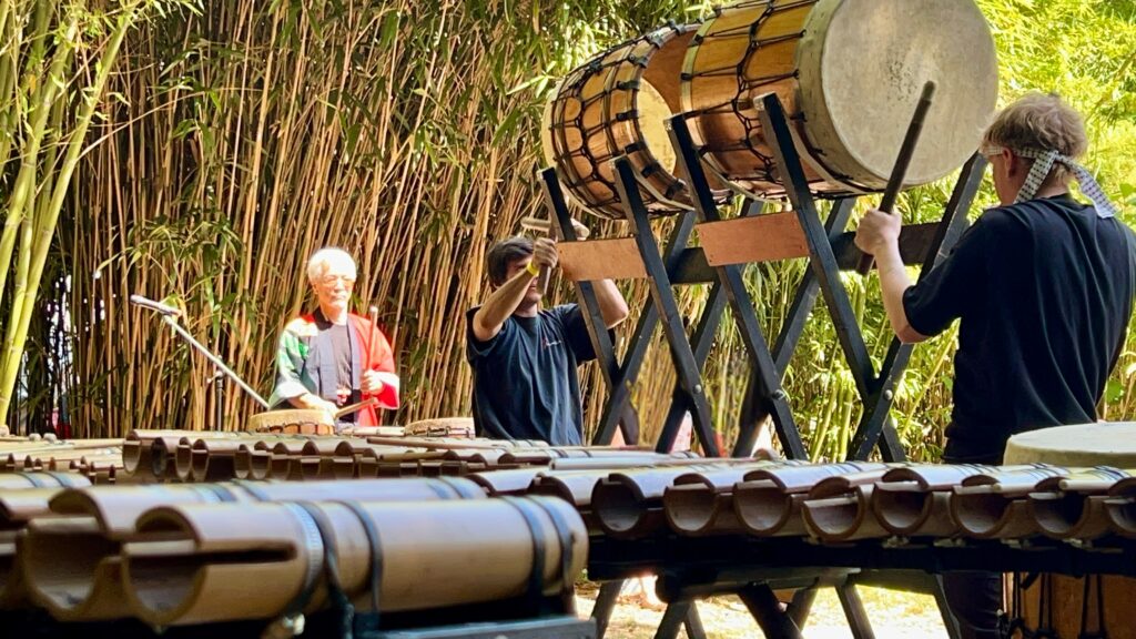Concert dans une bambouseraie : un musicien en veste japonaise rouge et deux percussionnistes autour d’un grand tambour japonais, une rangée d’instruments en bambou au premier plan.
