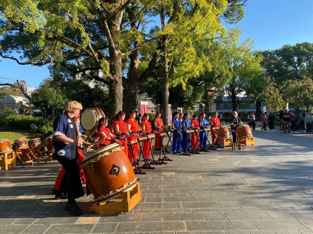 Une rangée de musiciens en tenue rouge joue des tambours dans un parc bordé d’arbres.
