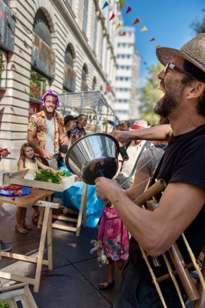 Un musicien en chapeau de paille, instruments en bambou en bandoulière, frappe une bassine métallique lors d’une animation de rue.