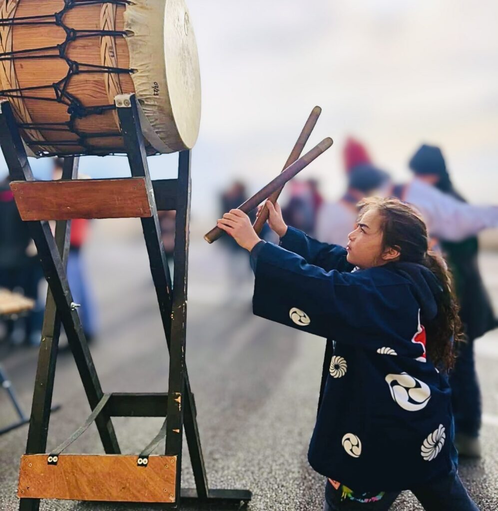 Une jeune musicienne en veste bleue traditionnelle frappe un grand tambour, baguettes levées.